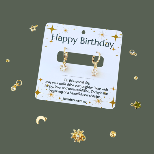 Gold earrings on a 'Happy Birthday' card with decorative elements on a dark green background