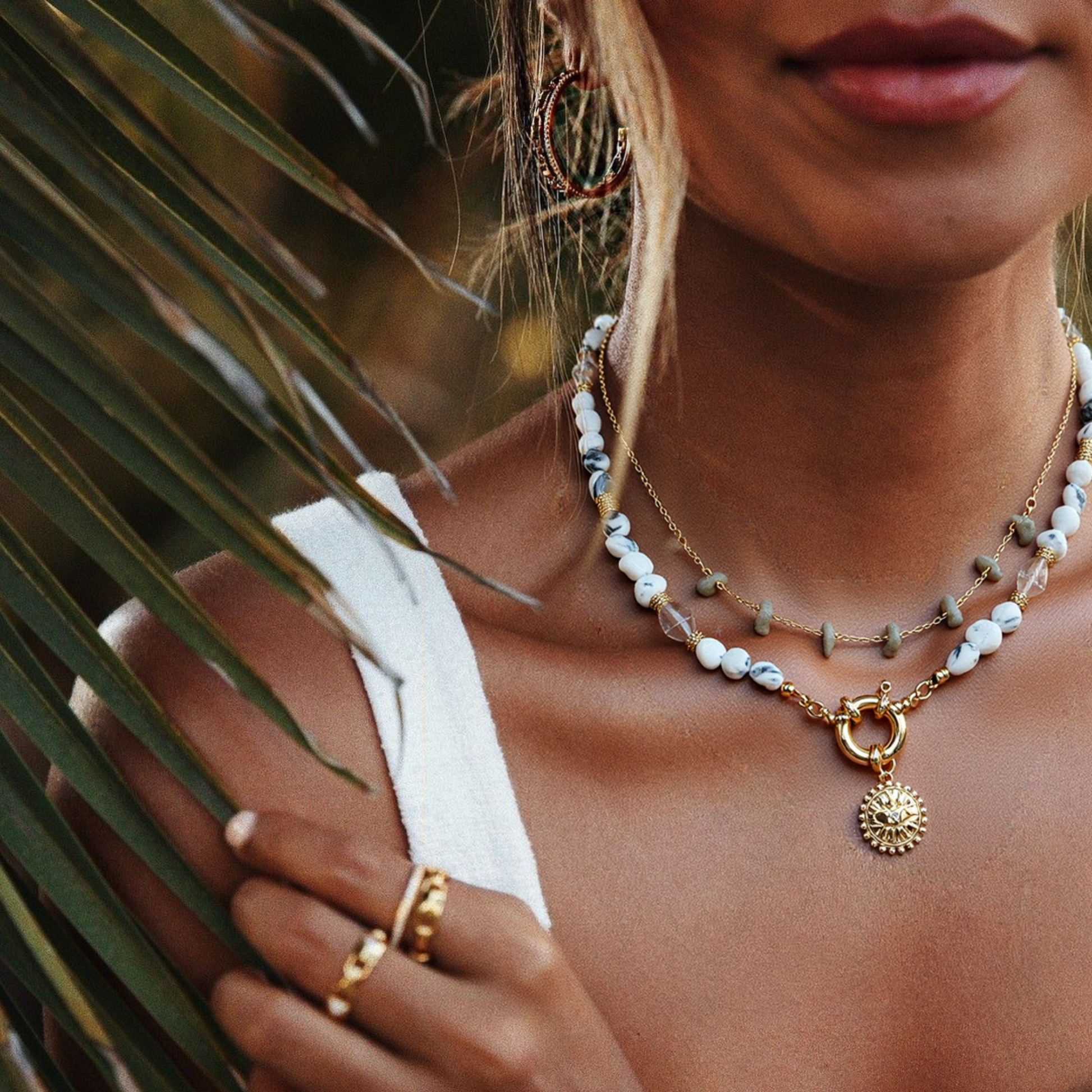 A woman wearing layered necklaces which including a nugget gemstone chain necklace and a natural howlite gemstone nacklace with Gold Plated Sunburst Evil Eye Pendant and holding a palm leaf