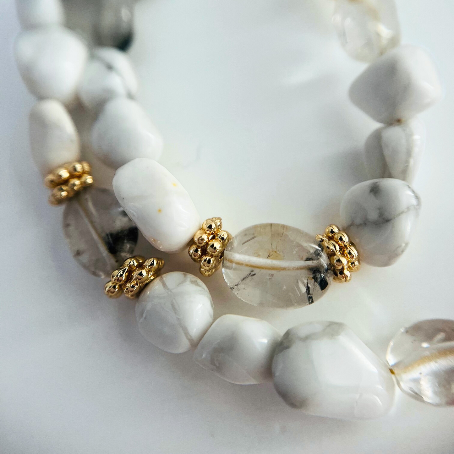 Close-up of a necklace with Howlite Nugget Gemstone and Black Rutilated Quartz, gold plated daisy spacer beads accents on a light background