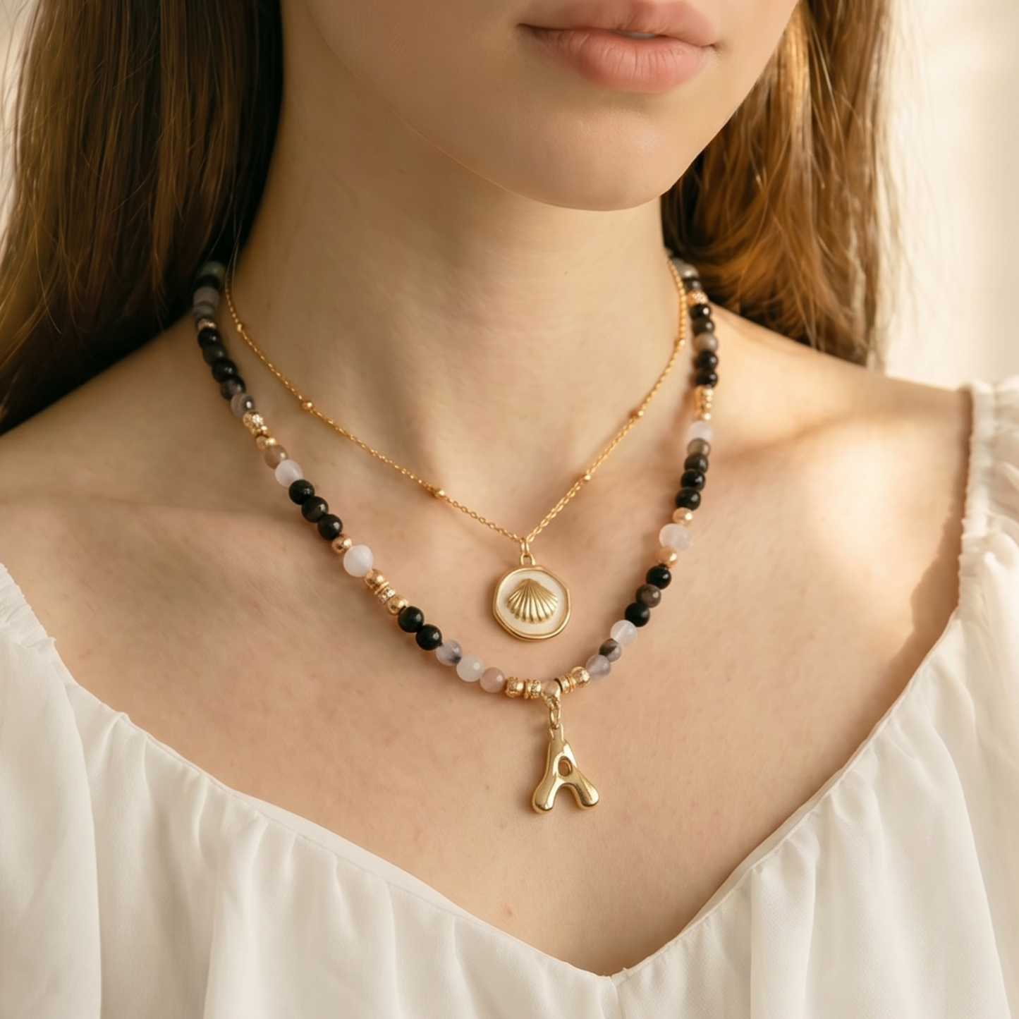 A woman wearing a delicate gold necklace with a small pendant and a beaded black and gold necklace, styled with a white ruffled top against a warm, softly lit background.